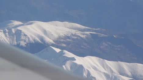 zoomed out view of snow capped mountains while looking out plane window and flying over europe with jet wing in frame