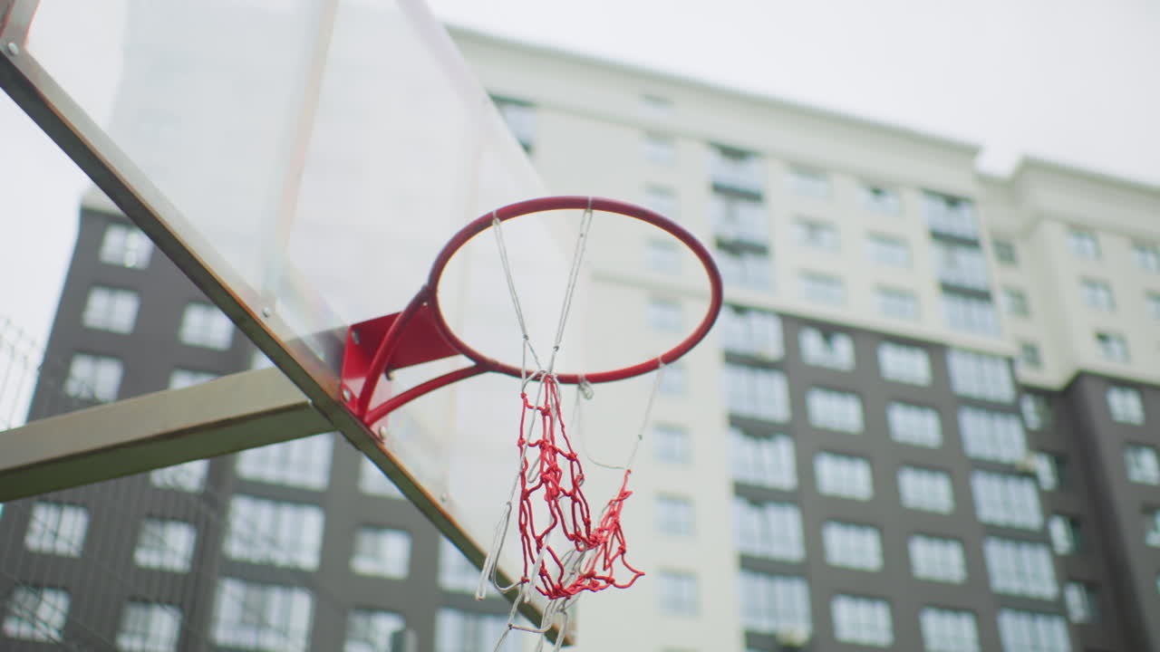 Canestro da basket urbano, primo piano, cerchio rosso, tabellone trasparente con rete a catena, grattacieli sullo sfondo, cielo nuvoloso, prospettiva dal basso, traiettoria della palla che suggerisce movimento, vernice usurata