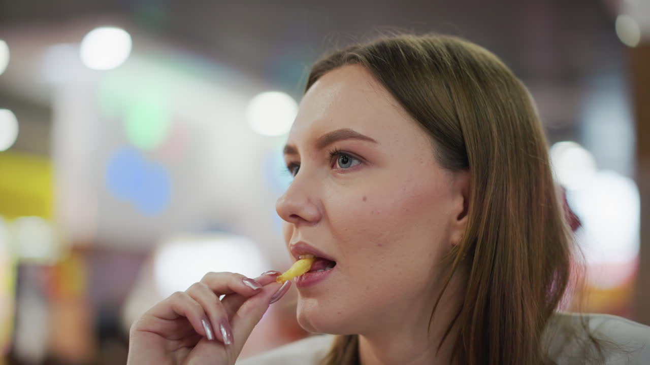 primer plano de una mujer comiendo papas fritas con una mirada enfocada, fondo bokeh suave con luces intermitentes coloridas en un entorno animado de un centro comercial