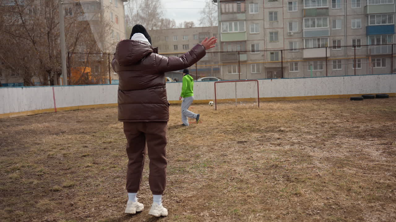 도심 안뜰에서 아이의 축구 킥을 코칭하는 백인 부모, 아파트 블록 배경, 격려를 몸짓으로 표현하는 갈색  пуховик 코트를 입은 부모, 드리블 및 슛을 하는 초록색 후드티를 입은 아이, 흐린 겨울 날씨
