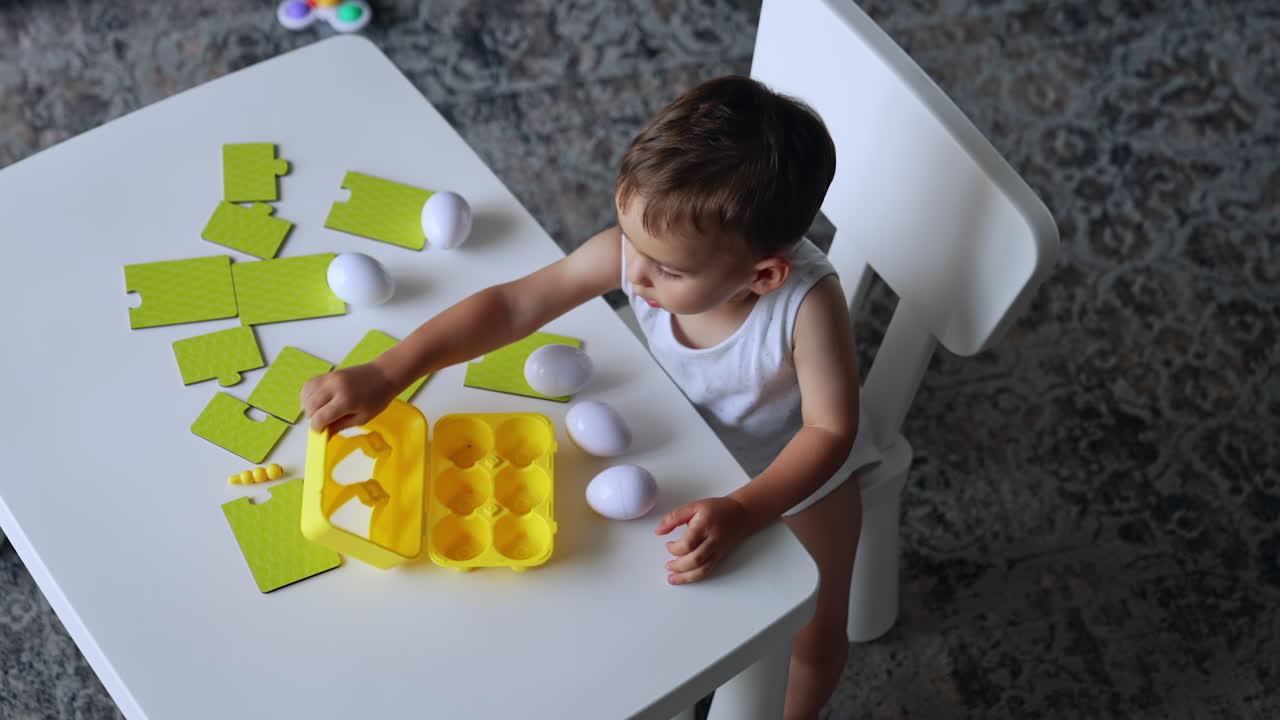 Lovely toddler boy sits at white desk. Cute child plays with toy eggs and puzzles. Top view.
