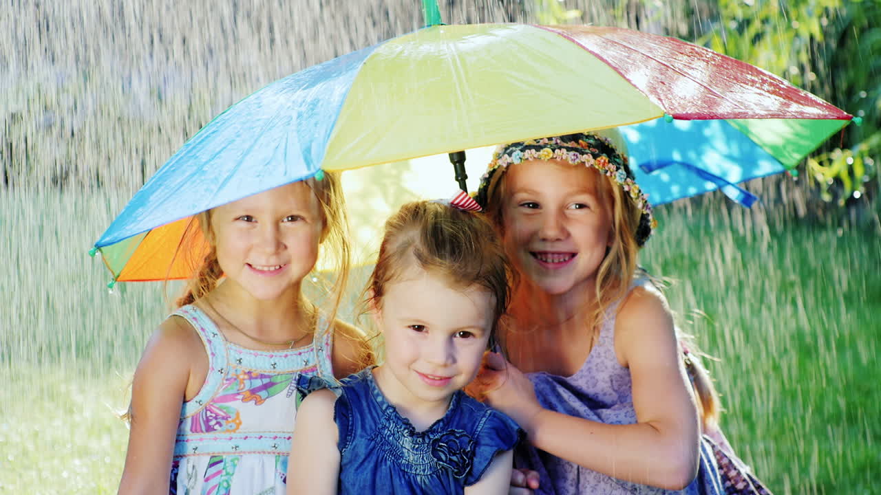 세 자매가 색상의 우산 아래 숨어 있습니다. 따뜻한 여름비가 내리고 있습니다. 아이들은 하