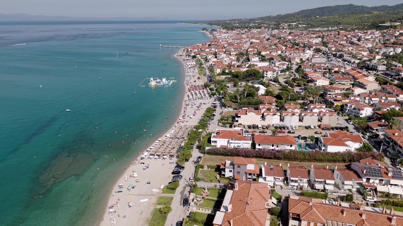 Spiaggia affollata di Pefkochori in estate, ville della nuova città del villaggio di Pefkochori, hotel, paesaggio montano e forestale sullo sfondo, Calcidica, Mar Egeo, penisola di Kassandra. Ripresa con drone dall'alto
