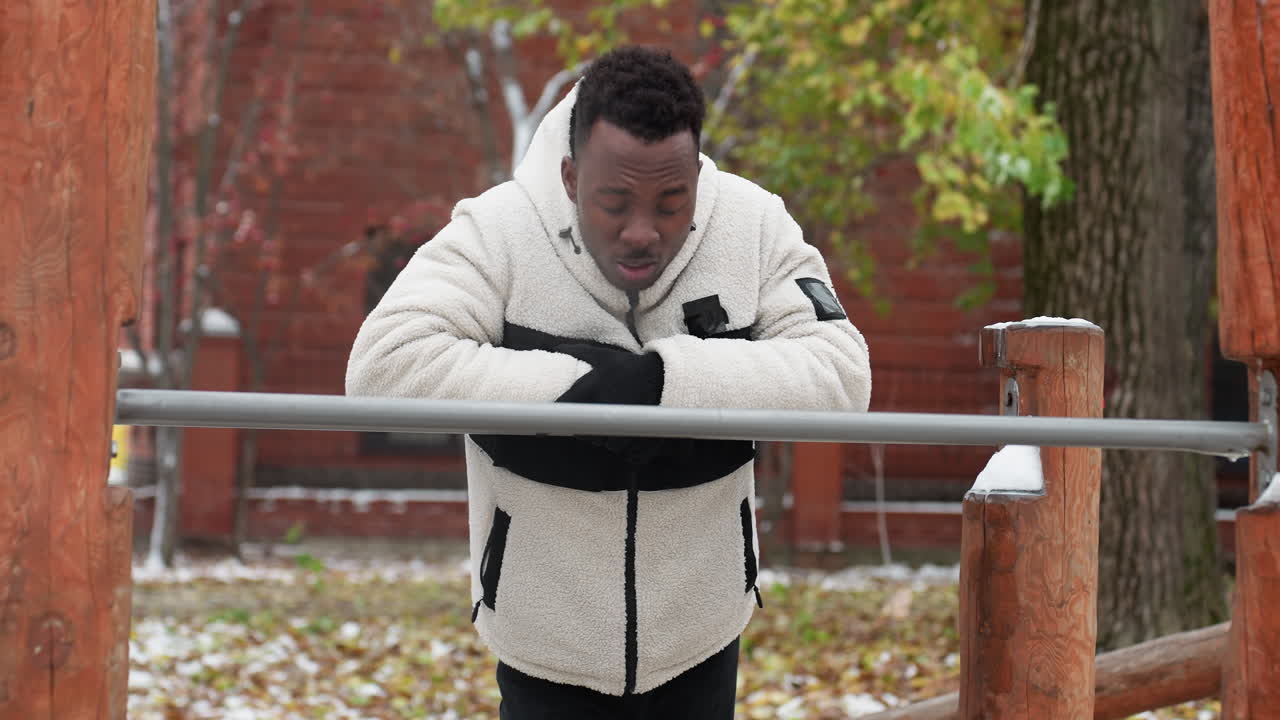Man resting on iron bar after intense workout session in cold weather, dressed in a warm fleece jacket, he catches his breath while leaning forward, snow-covered ground and trees in background