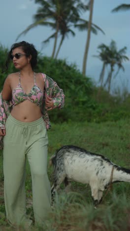 woman in tropical outfit stands confidently in lush field with goat, palm trees in background