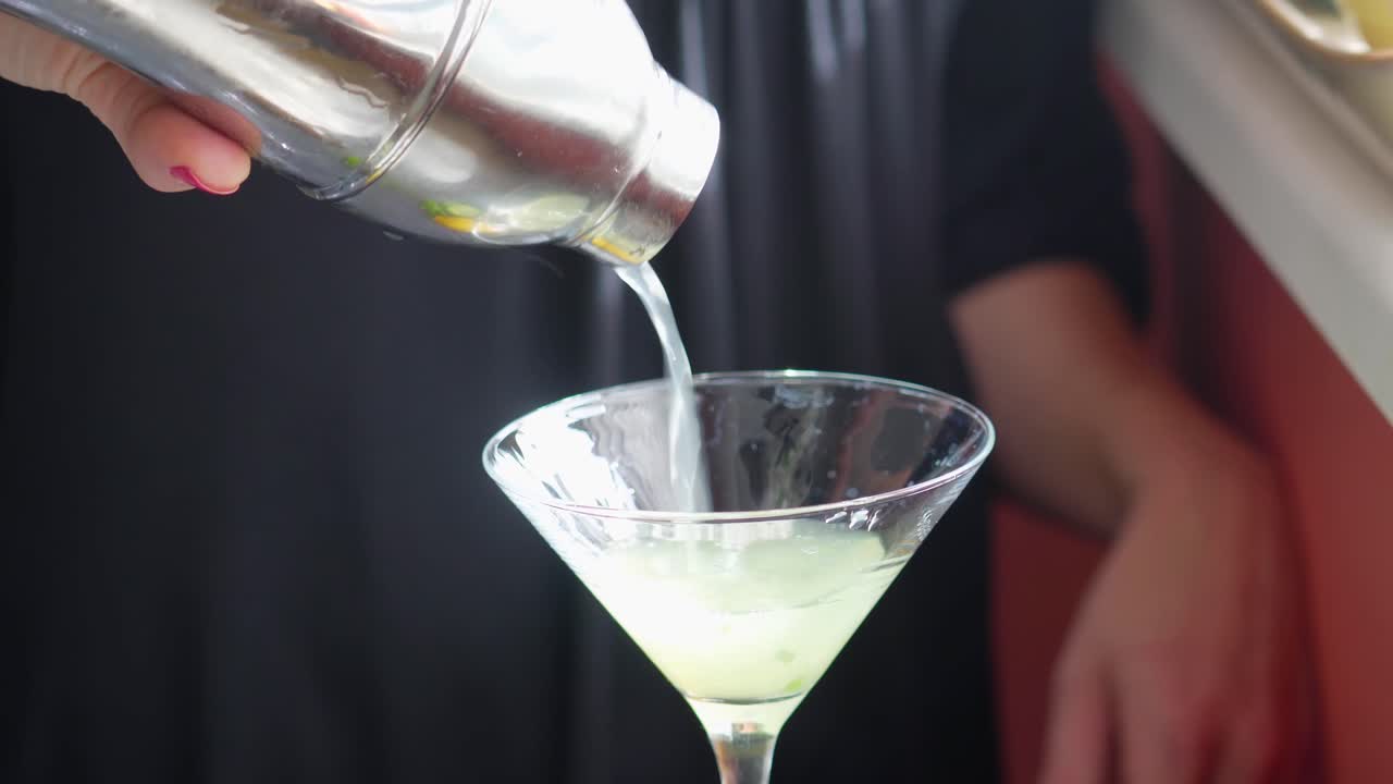 el camarero vierte un cóctel o una bebida sin alcohol en un vaso de martini de una coctelera, hojas de menta verde y una bebida mixta de pepino gotea lentamente de la mano de una camarera, alcohol