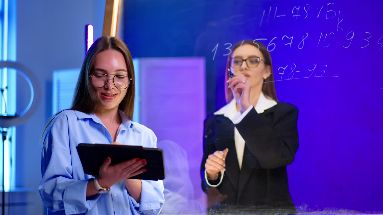 Classroom of young women. Two young women engage in a classroom discussion. One holds a tablet while the other explains on a transparent board