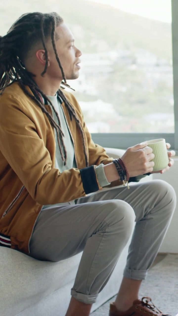Vertical video: Prompted by window light in living room, man in bomber sipping green mug, gesturing