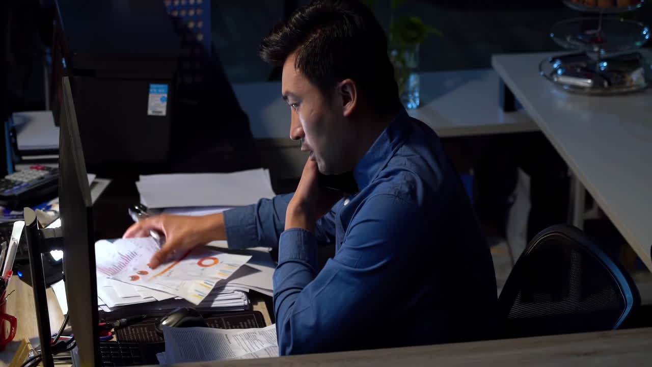Young businessman feeling overwhelmed by a mountain of paperwork. He is feeling stressed and frustrated as he looks at computer screen