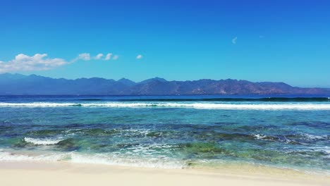 Paisaje-Marino-Pacífico-Con-Olas-Blancas-De-Mar-Que-Cubren-El-Lecho-Marino-Rocoso-Cerca-De-La-Playa-De-Arena-Blanca-En-Un-Cielo-Azul-Brillante-Sobre-El-Horizonte-De-Las-Montañas-En-Bali