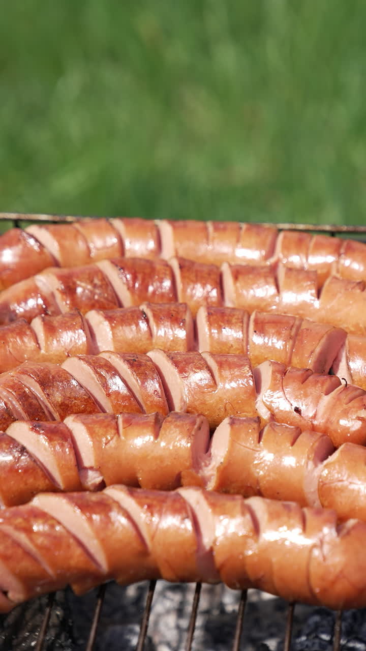 Tasty fried sausages on a grill. Grilled sausages with incisions ready to eat for a picnic. Hot dog sausages roasting in charcoals outdoors. Barbeque. Vertical video