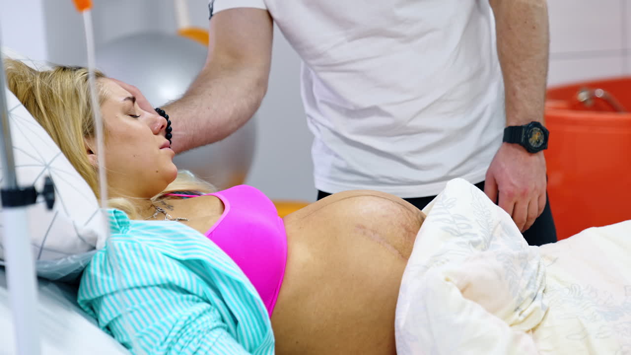 Pregnant Caucasian woman suffers from contractions. Lady lies in bed and her husband stands behind stroking her head.