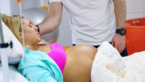 pregnant caucasian woman suffers from contractions. lady lies in bed and her husband stands behind stroking her head.