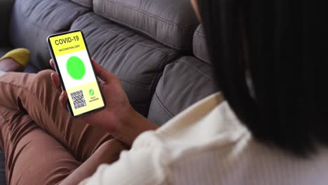 Woman-at-home-holding-smartphone-with-covid-vaccination-certificate-and-qr-code-on-screen