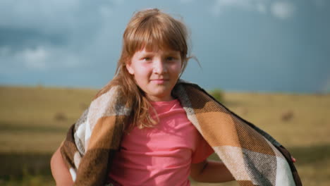 una niña confiada con una camiseta rosa caminando con gracia a través de un vasto campo abierto con una manta sobre los hombros, el viento soplando, balas de heno doradas en la distancia