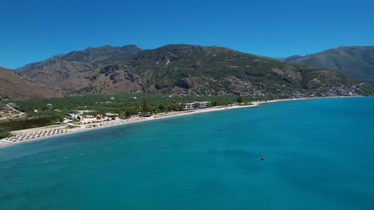 Bay with Azure Seawater on Ionian Coastline of Albania, Calm Beach with White Sand, Green Olive Grove, and Mountains in Background