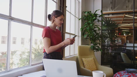 Young-Hispanic-woman-standing-by-the-window-in-a-creative-office-using-smartphone,-panning-shot