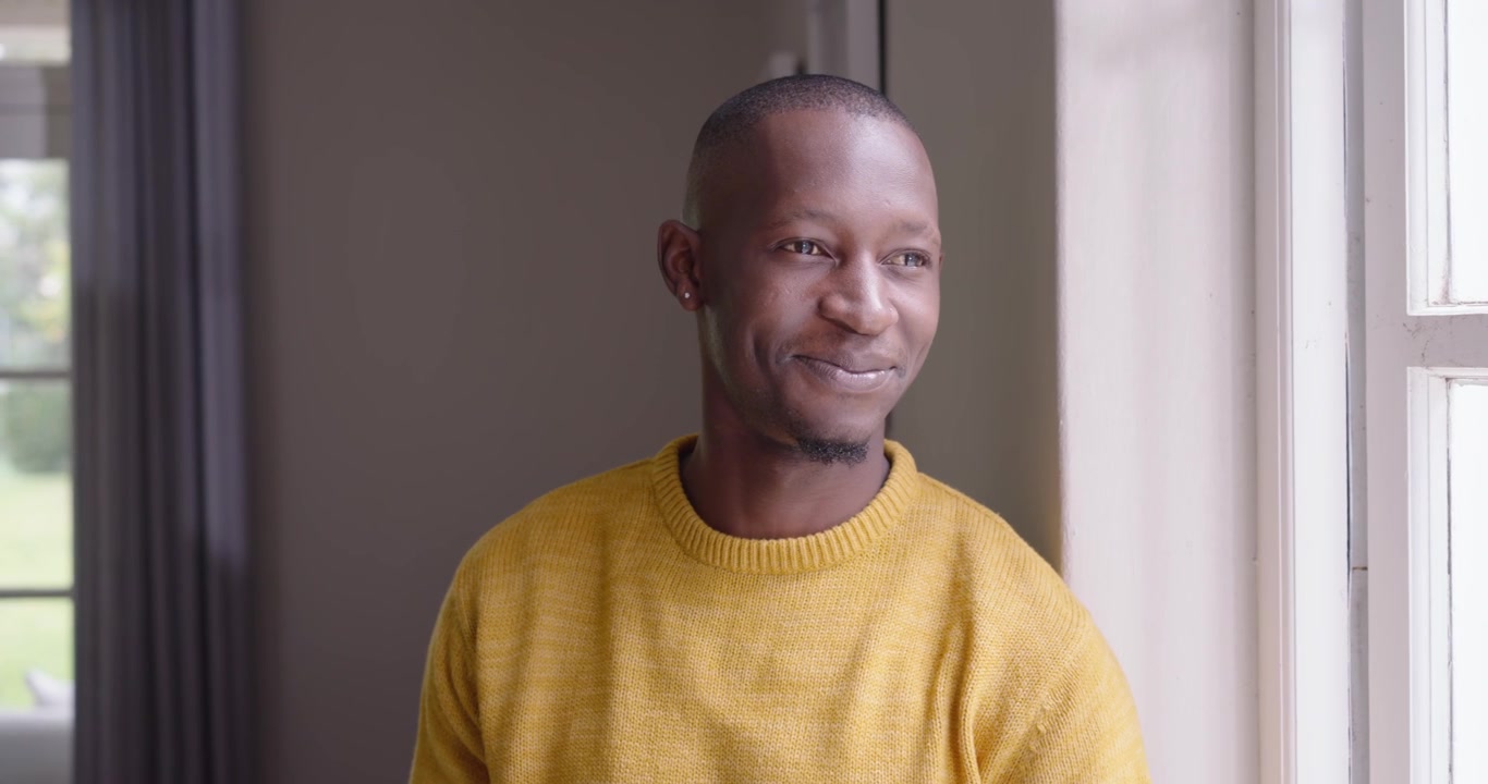 Homme africain d'une vingtaine d'années, assis près d'une grande fenêtre, remarquant une voix hors champ, tournant la tête et souriant.