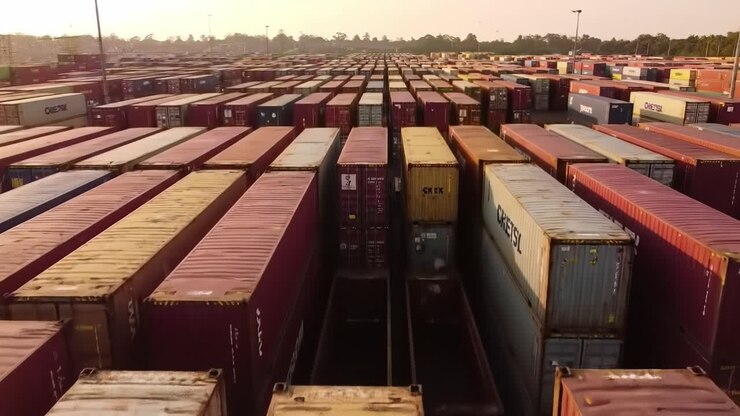 Witness a sprawling shipping container yard capturing an expansive aerial view. Containers are meticulously arranged, showcasing brilliant colors as the sun sets in the background.