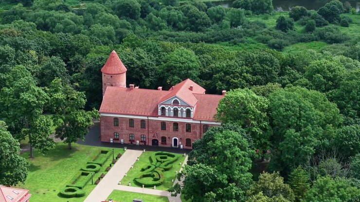 Drone video captures Raudondvaris Castle’s red brick towers, manicured gardens, and lush green parkland in vibrant daylight, highlighting historical architecture and scenic Lithuanian landscape.