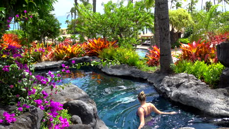 Young-blonde-woman-wades-through-colorful-resort-pool-at-Poipu-Beach-on-the-garden-Isle-of-Kauai,-Hawaii,-4k,-with-model-release