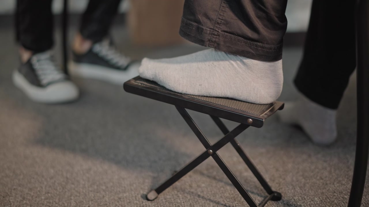 Voet met sok op een voetenbankje tijdens een gitaarles; close-up van een student die een opvouwbare voetensteun aanpast terwijl de leraar in sneakers toekijkt, met zachte vloerbedekking, warm natuurlijk licht en gerichte, geduldige techniekcoaching.
