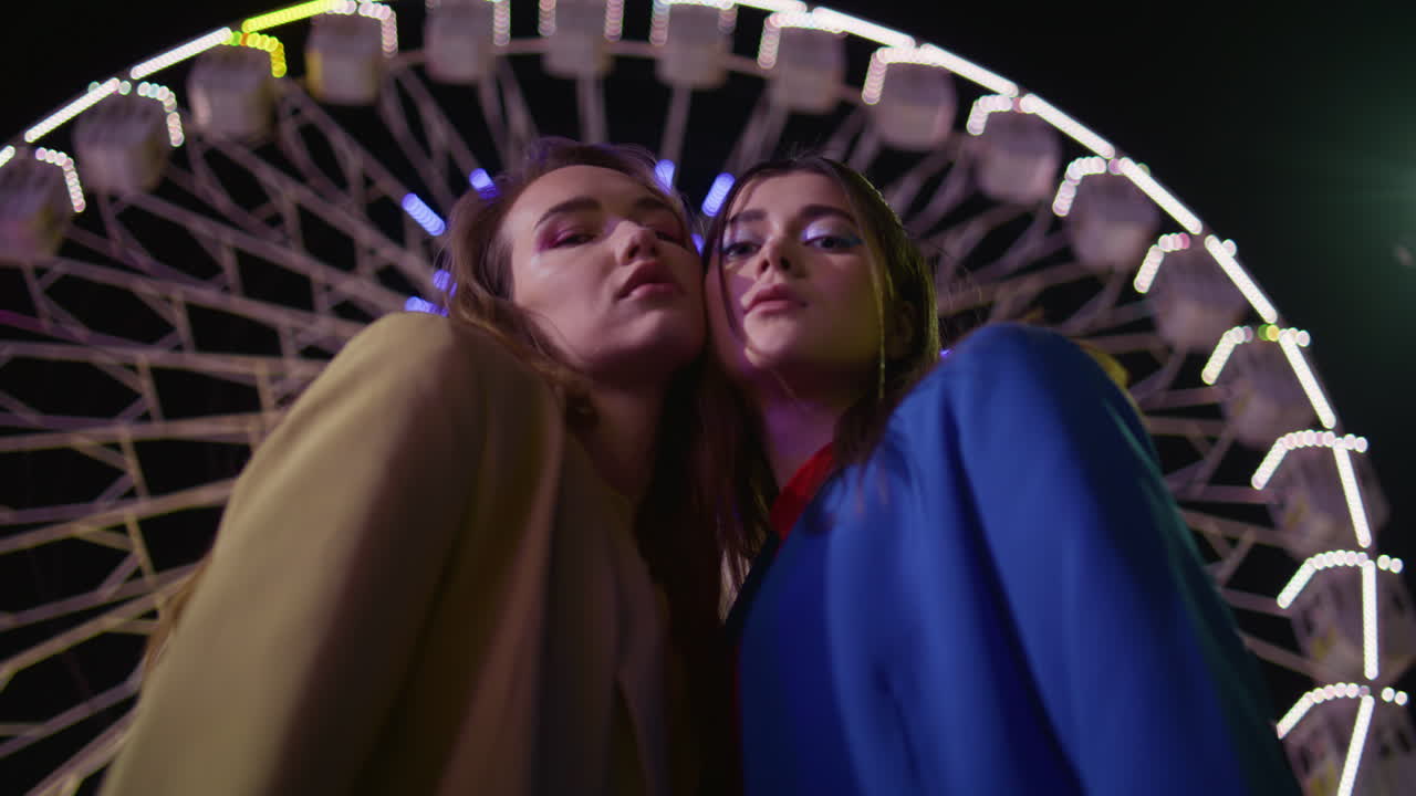 Closeup girls couple posing at blinking ferris wheel. Fashion models look camera
