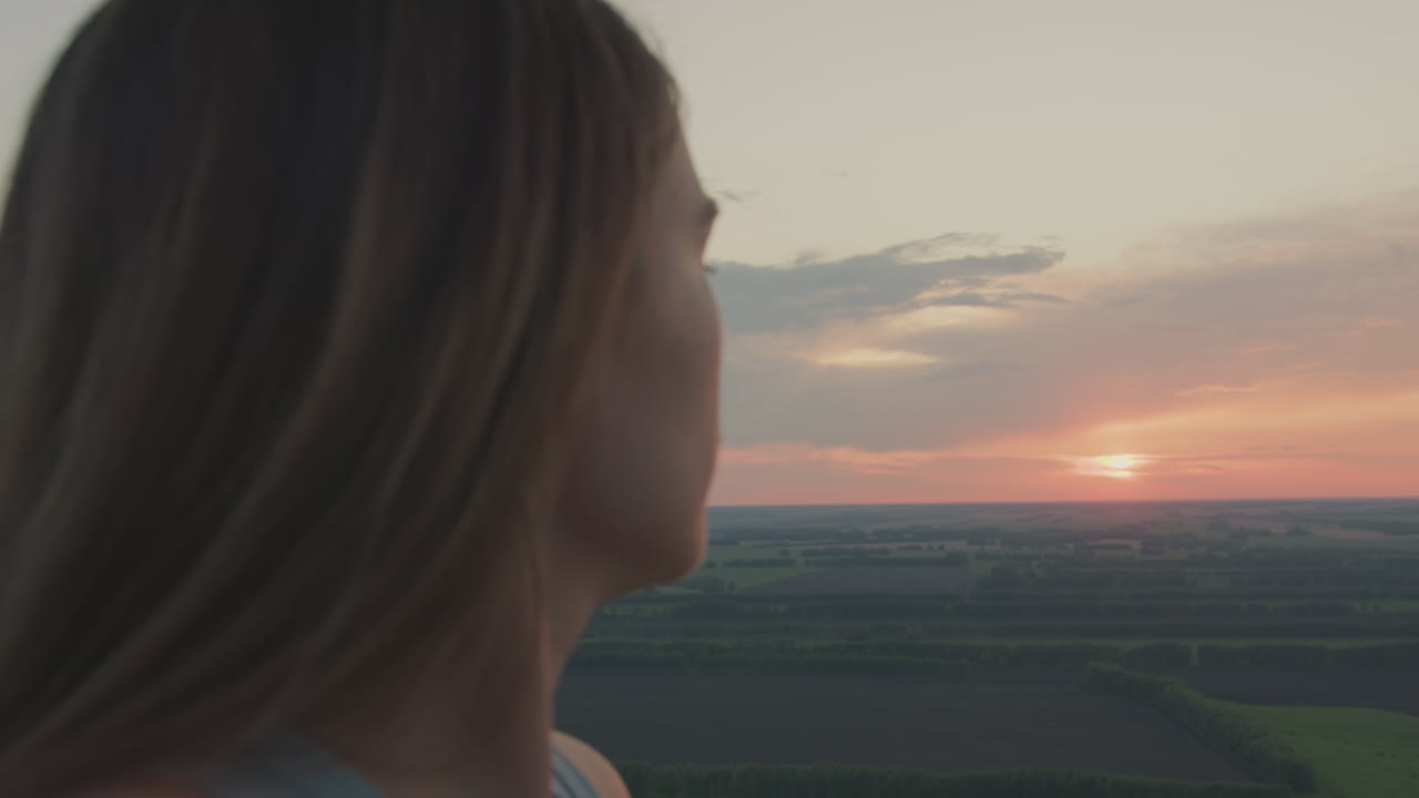 Lady gazes across glowing sunrise horizon from hot air balloon basket high above vast patchwork fields, morning light illuminating profile as serene sky and rolling landscape stretch into distance