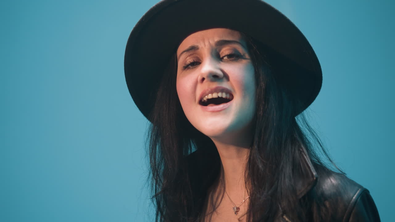 primer plano de una dama con un sombrero negro y una chaqueta de cuero negra, cantando apasionadamente con las manos levantadas contra un fondo azul vibrante. su cara expresiva