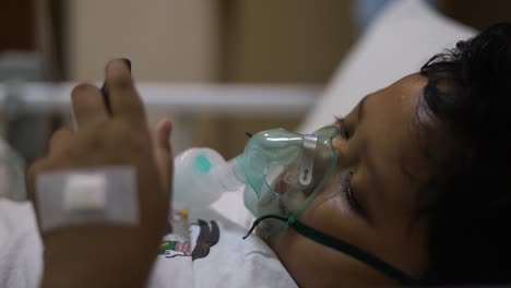 lindo niño pequeño con máscara de oxígeno en la cara en la cama en el hospital mientras juega al teléfono celular