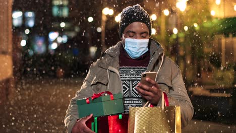 portrait of young african american man wearing facial mask typing on smartphone and holding shopping bags on the street while it¬¥s snowing in christmas