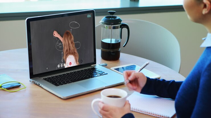 Estudiante caucásica usando una laptop en videollamada con una profesora, tomando apuntes.