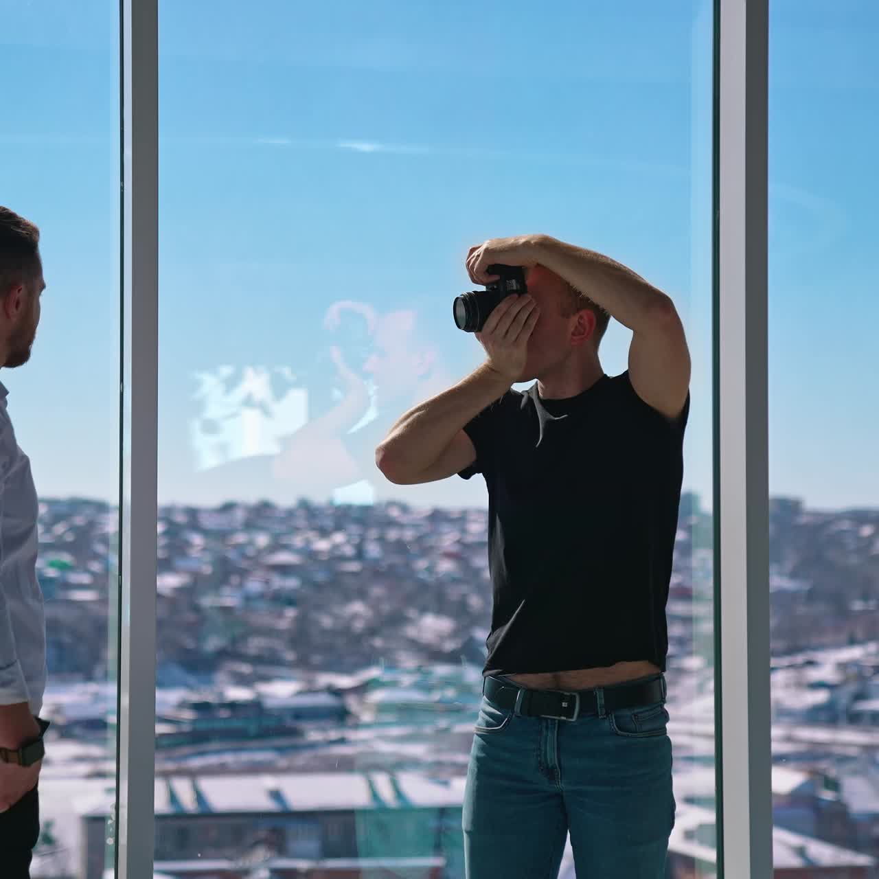 Young photographer taking photos of a businessman. Man shooting handsome man model in white shirt near windows. Working with camera. Profile view
