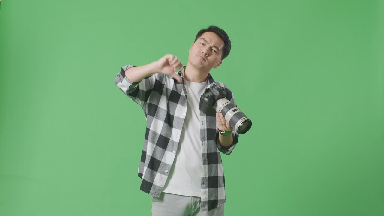 화가 난 아시아 사진작가는 카메라에서 사진을 보고 스튜디오에서 녹색 화면 배경에 서 있는 동안 엄지손가락을 아래로 표시합니다.