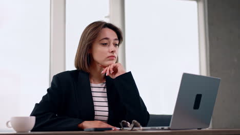 thoughtful young lady sits at desk. woman with dark hair wearing black jacket looks at laptop.