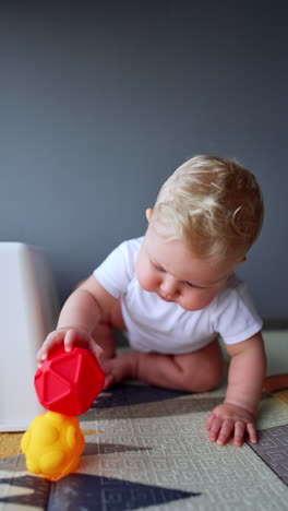 adorable baby boy wearing white bodysuit sits on the floor. cute blond toddler plays with his toys at home. vertical video.
