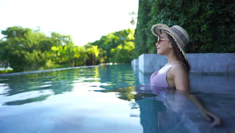 Vista-Lateral-De-Una-Mujer-Asiática-Sentada-En-Una-Piscina-Infinita-Con-Sombrero