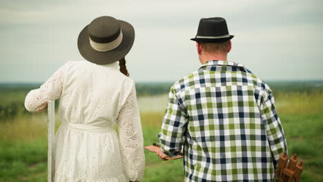 Close-up back shot of a couple walking on a grassy field. The woman, dressed in a white gown, carries a canvas while the man, in a checkered shirt and jeans, hands her a drawing pad as they