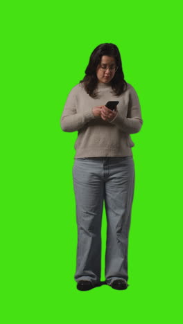 Vertical-Video-Full-Length-Shot-Of-Young-Woman-Checking-Text-Message-On-Mobile-Phone-Standing-Against-Green-Screen