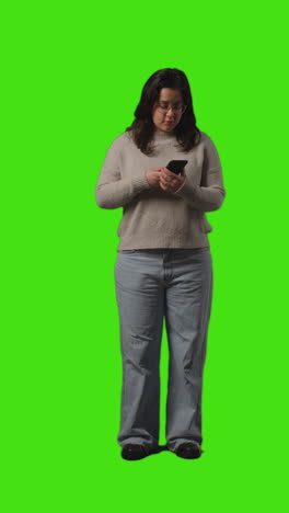 vertical video full length shot of young woman checking text message on mobile phone standing against green screen