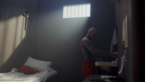 tattooed inmate washing his hands and face above sink in prison cell