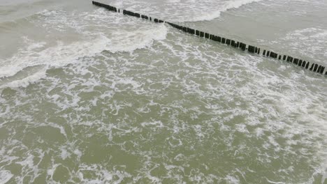 Aerial-birdseye-view-of-Baltic-sea-coast-on-a-overcast-day,-old-wooden-pier,-white-sand-beach,-large-storm-waves-crushing-against-the-coast,-climate-changes,-wide-drone-shot-moving-forward