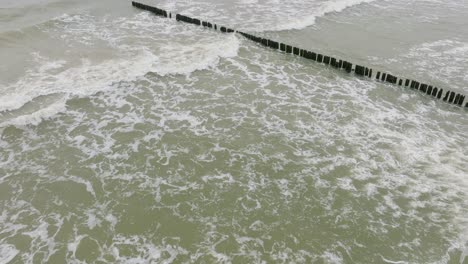 vista aérea de pájaro de la costa del mar báltico en un día nublado, antiguo muelle de madera, playa de arena blanca, grandes olas de tormenta aplastando la costa, cambios climáticos, gran tiro de drones avanzando