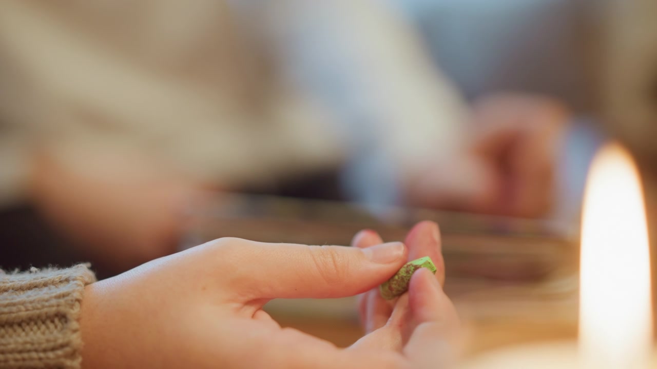 Close up of hand holding and examining green crayon with soft lighting. Background shows another person working at table creating warm indoor scene with candlelight