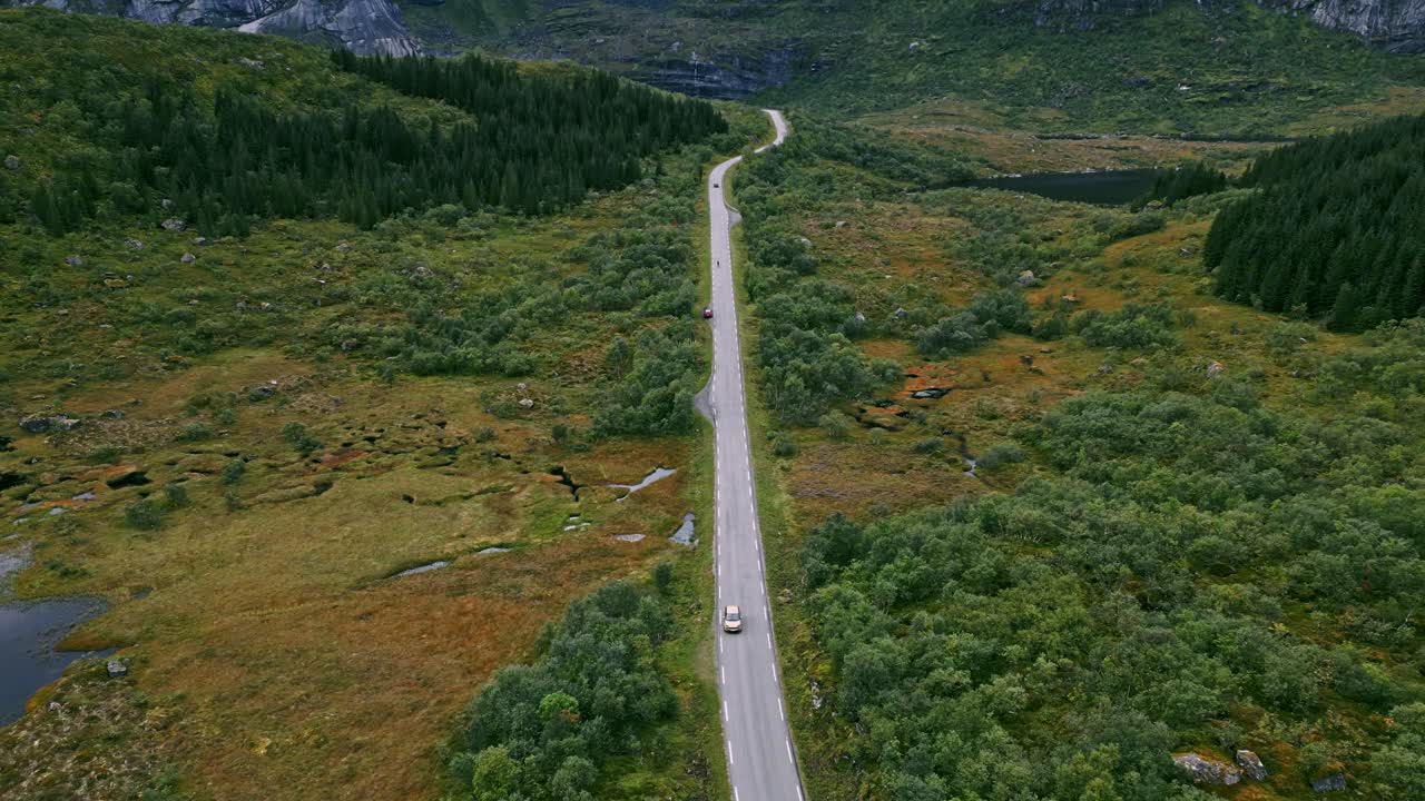 toma panorámica frente a un automóvil dorado que conduce a lo largo de la carretera escénica en lofoten, noruega, que revela un paisaje panorámico de montañas y lagos espectaculares