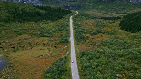 Schwenkflugaufnahme-Vor-Einem-Goldenen-Auto,-Das-Entlang-Der-Malerischen-Straße-Auf-Den-Lofoten-In-Norwegen-Fährt-Und-Eine-Panoramalandschaft-Mit-Dramatischen-Bergen-Und-Seen-Offenbart