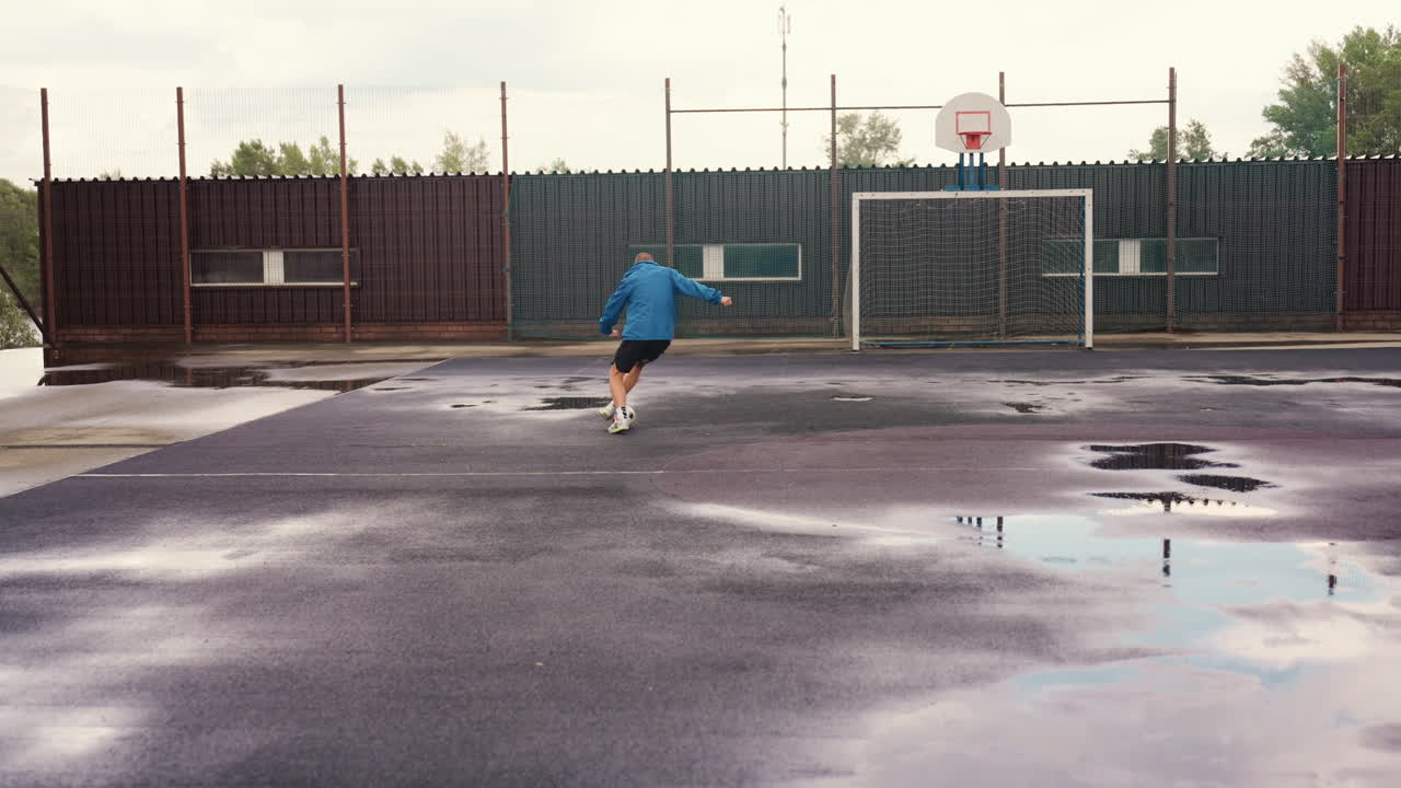 Campo di asfalto zuppo di pioggia, giocatore che si allena da solo a calcio palleggiando vicino a pozzanghere e alla porta, giacca blu, scarpe da ginnastica, schizzi, cielo nuvoloso, sfondo di recinzione urbana, allenamento mirato, movimento dinamico