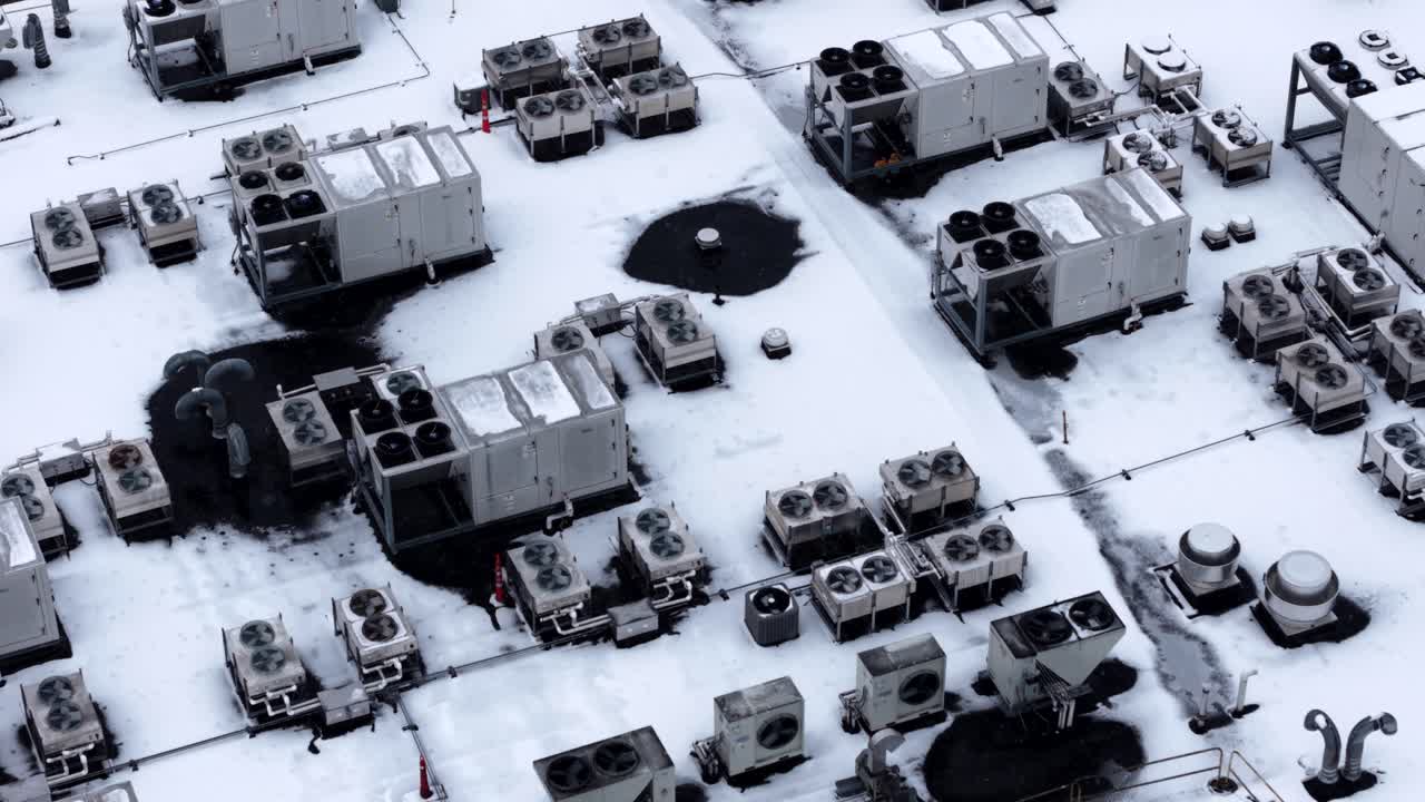 Aerial winter view of an American commercial rooftop covered in snow, with multiple HVAC units, ventilation systems and industrial equipment arranged in grid under cold conditions. Top down