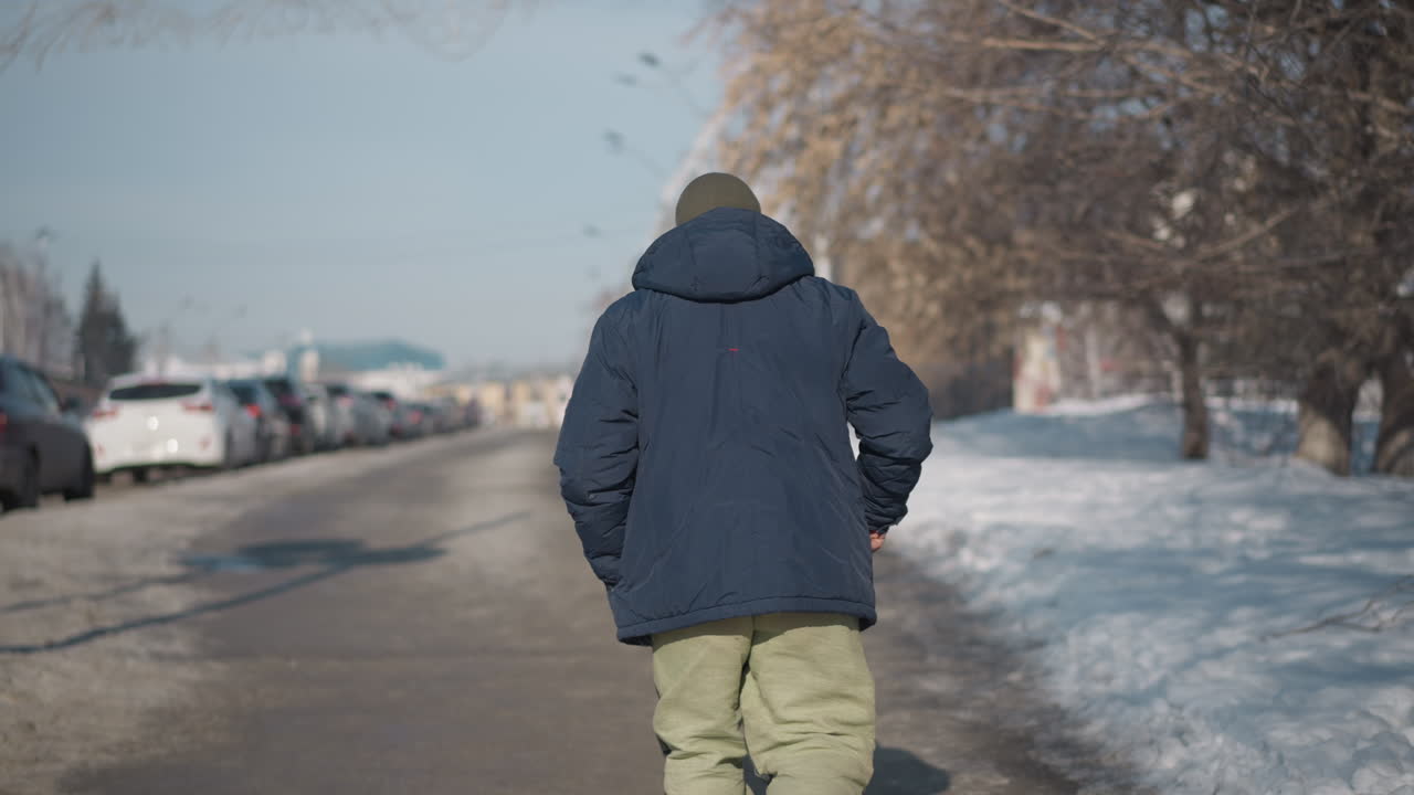 back view young man wearing black jacket strolling along sunlit snow covered street with parked cars lining pathway, footprints in slush, winter trees and buildings blurred in background
