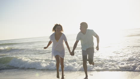 una pareja biracial disfruta de una carrera lúdica a lo largo de la playa al atardecer